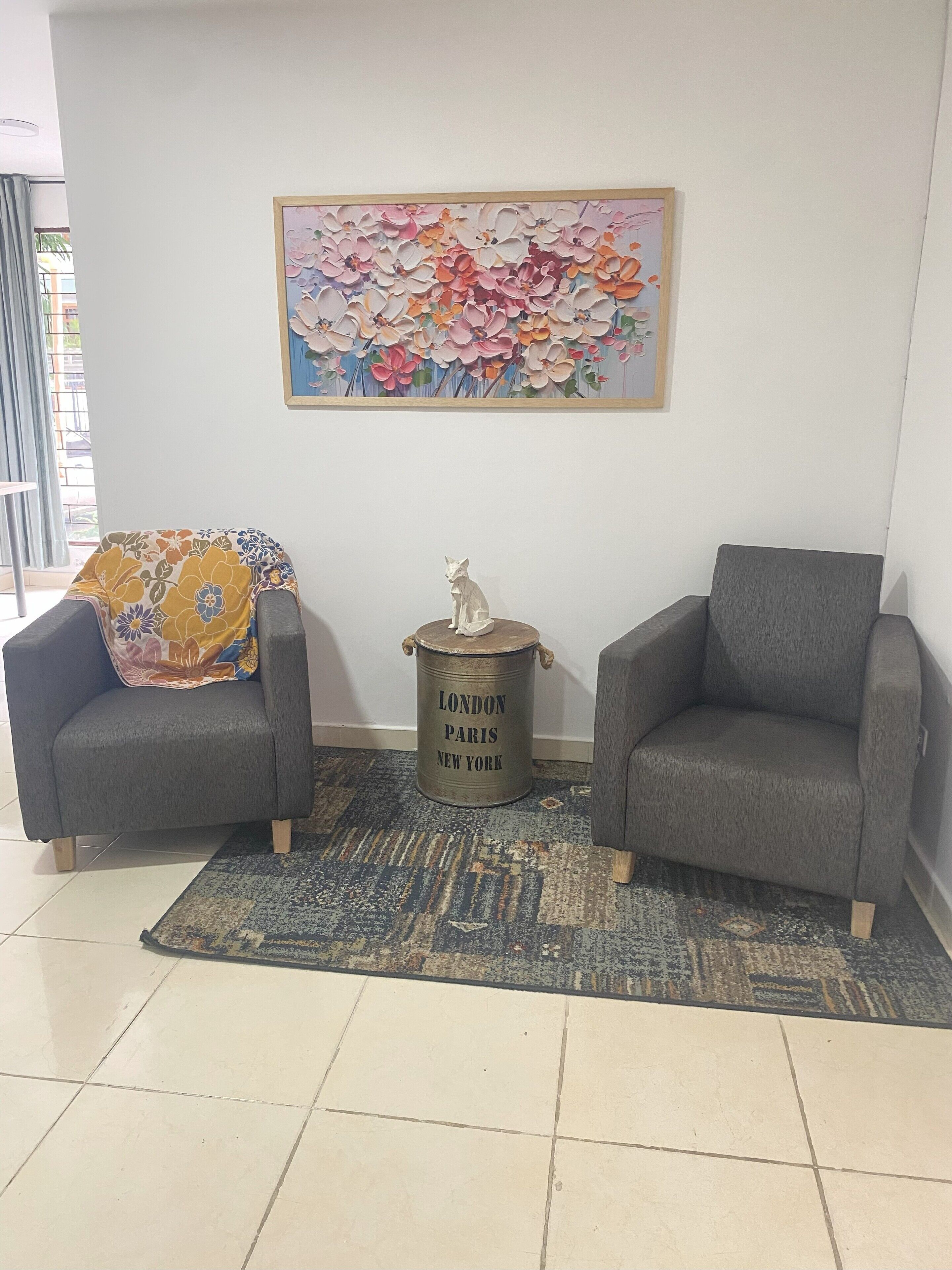Een kamer met twee grijze fauteuils, een geborduurd plaid, een decoratieve metalen vaas en een bloemschilderij aan de muur.