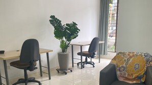 Una habitación con dos escritorios, sillas, una planta en maceta y un sofá con un cojín floral.