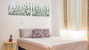 Un dormitorio con cama, una mesita con una planta y un cuadro floral en la pared.