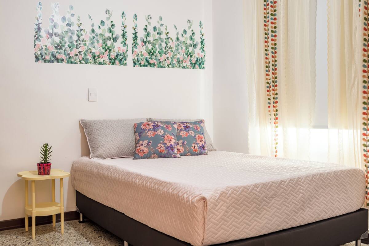 Un dormitorio con cama, una mesita con una planta y un cuadro floral en la pared.