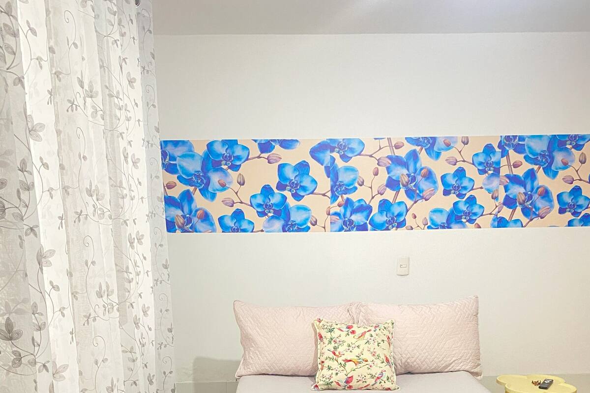 Un dormitorio con cama, un cojín estampado con motivos florales, una mesita de noche pequeña con un teléfono, y una pared cubierta de papel tapiz floral azul.