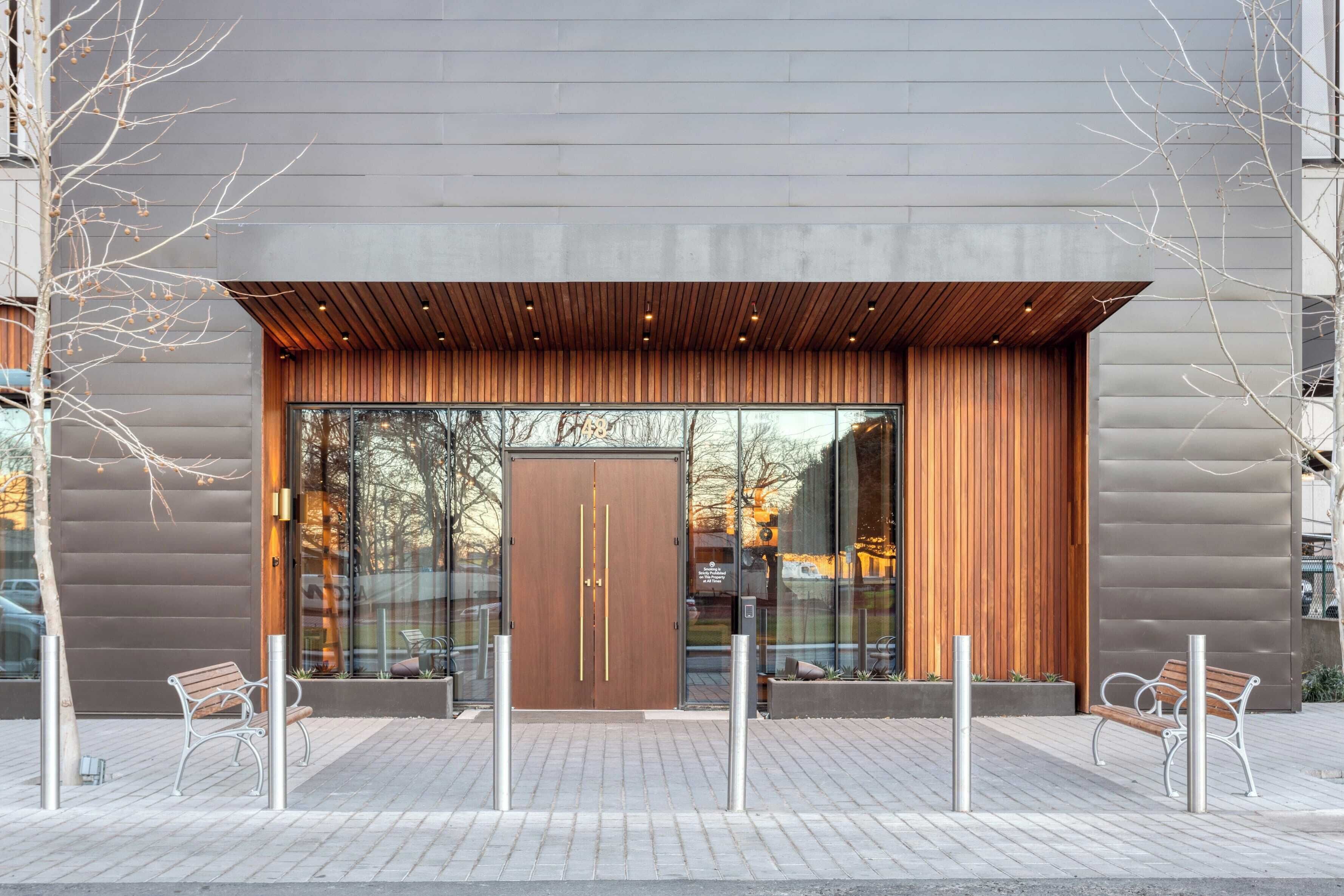Entrada de un edificio moderno con una puerta de madera grande, pilares metálicos y dos bancos en la acera.