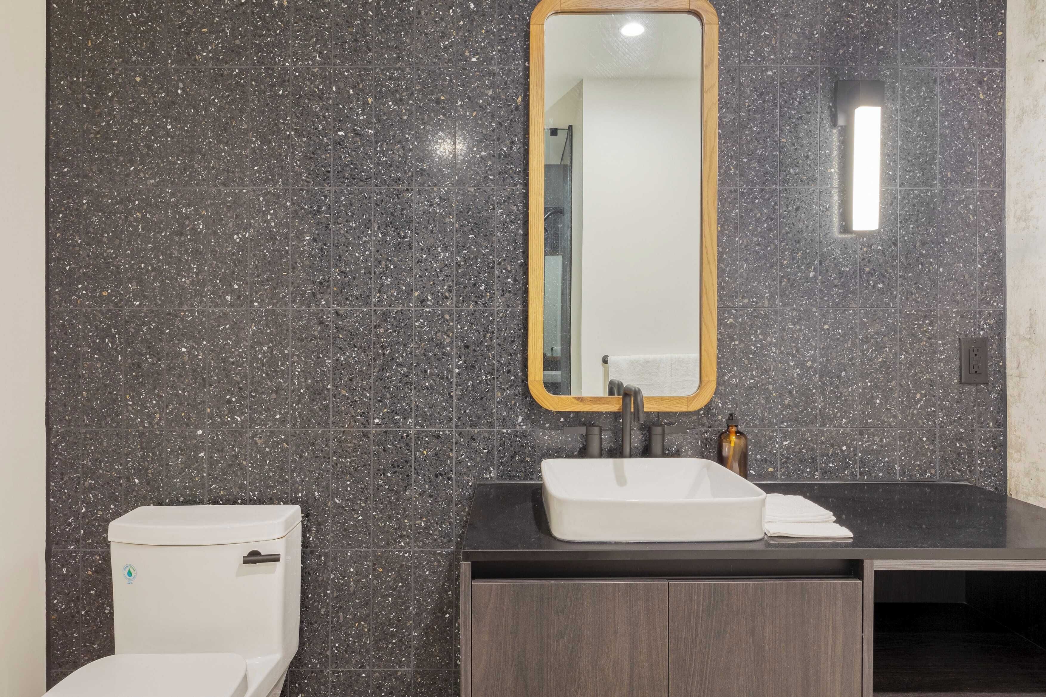 A bathroom with a white toilet, a rectangular mirror with a wooden frame, a black countertop, and a white sink.