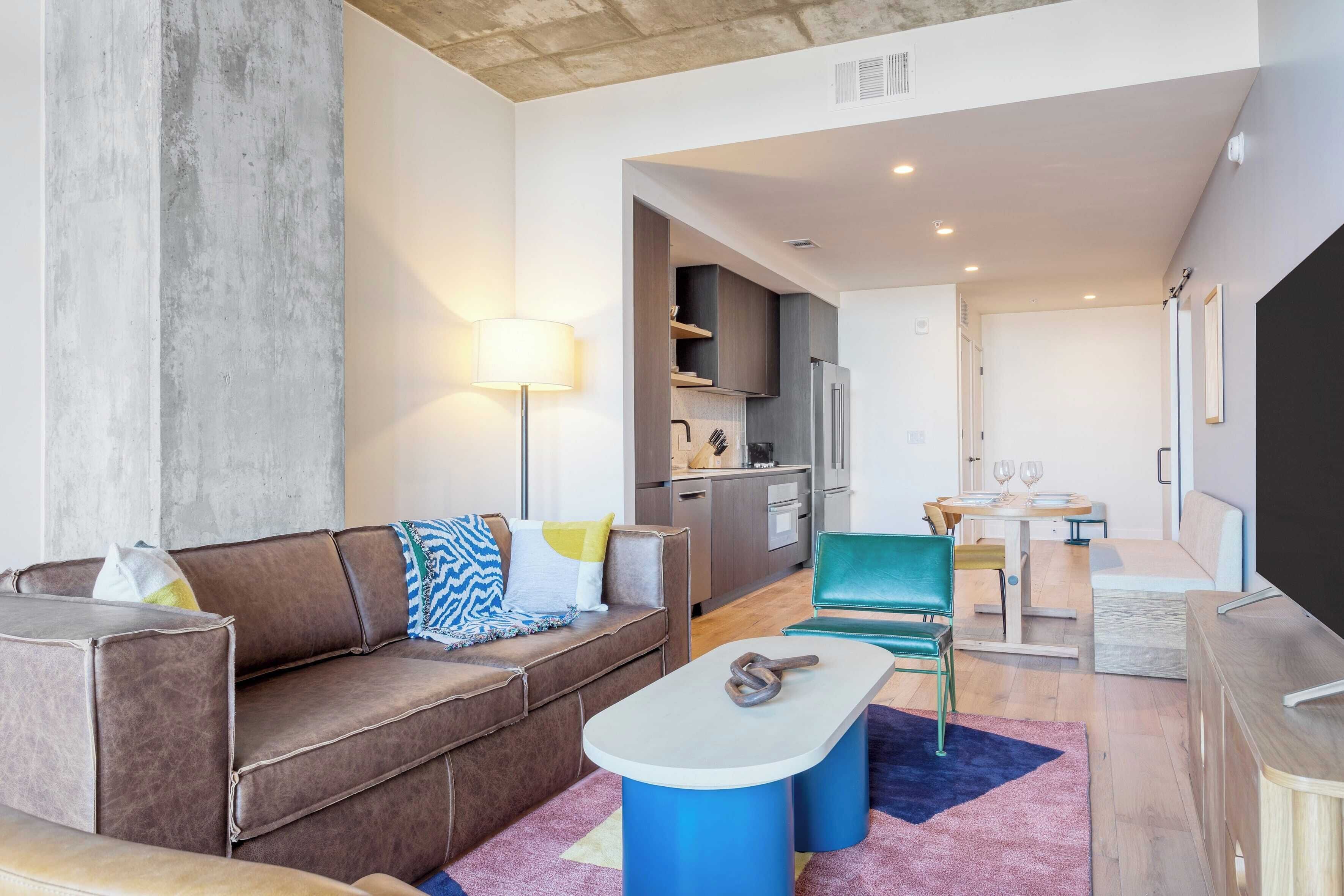 A modern living room with a brown leather sofa, a blue coffee table, and a dining area with a green chair and a small table.
