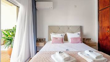 Habitación de hotel con cama, dos mesitas de noche, cabecero, unidad de aire acondicionado, una puerta y una planta en maceta.