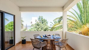 Une terrasse ensoleillée avec une table ronde, des chaises en osier et une vue sur la mer et la verdure.