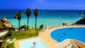 Une piscine où des gens nagent, une plage bordée de palmiers et un ciel bleu azur.