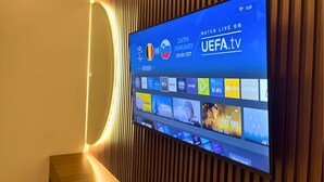 Televisão de tela plana fixada na parede, exibindo um evento esportivo e diversos serviços de streaming.