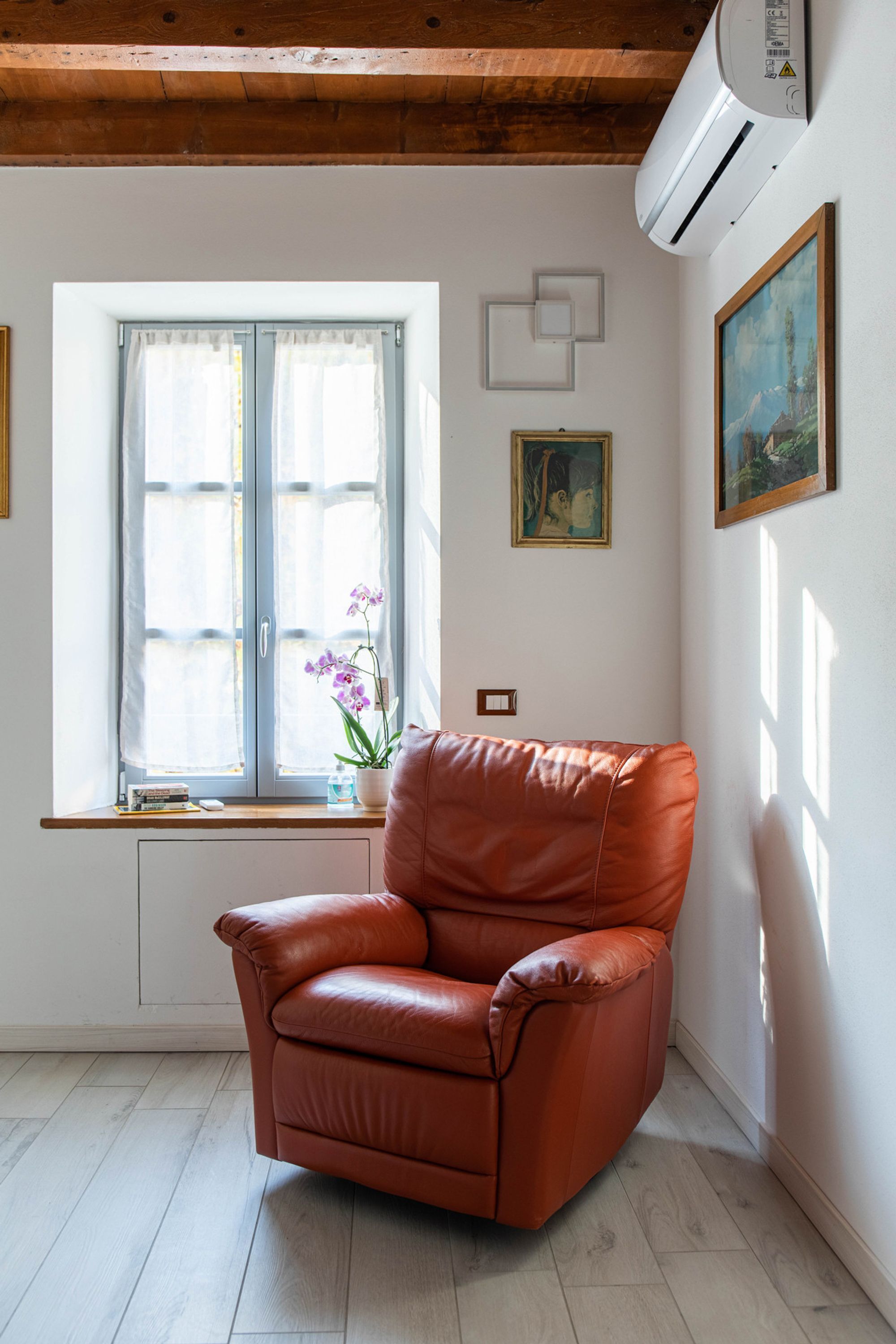 Une pièce avec un fauteuil en cuir marron, une fenêtre avec des rideaux, une orchidée en pot et un tableau accroché au mur.