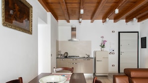 A dining area with a wooden table, chairs, and a bowl. A kitchen is visible in the background with a refrigerator, stove, and sink. - Varenna Vintage by Wonderful Italy (Perledo)