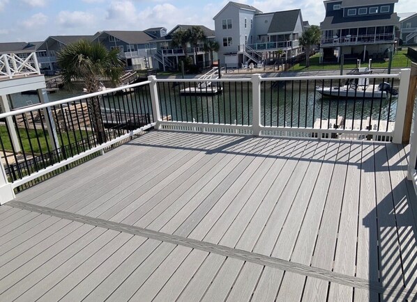 A wooden deck with a railing, overlooking a marina with boats and houses in the background. - NEWLY RENOVATED Canalfront w/ Boat Dock & Close to the Beach by Williamson Realt (Ocean Isle Beach)