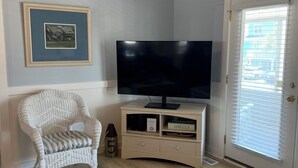 Ein Wohnzimmer mit Fernseher auf einem Schrank, einem Korbstuhl, einem gerahmten Bild an der Wand und einem Glastisch mit Meeresschalen.