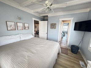Un dormitorio con una cama grande, ventilador de techo, televisor montado en la pared y puerta que da a otra habitación.