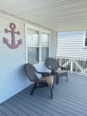 A covered porch with two wicker chairs, a large anchor decoration, and a window with blinds.