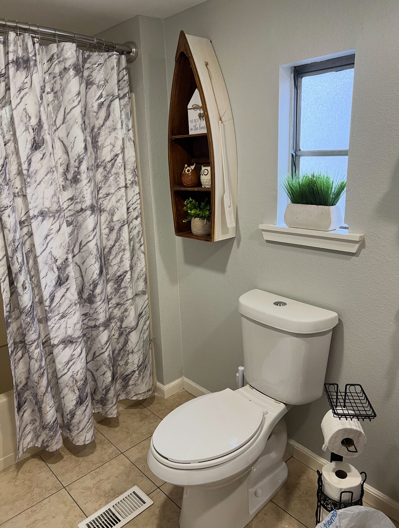 Ein Badezimmer mit Toilette, einer Duschvorhang, einem Ablagebereich mit Dekoration, einem Fenster mit Pflanze und einem Toilettenpapierhalter.