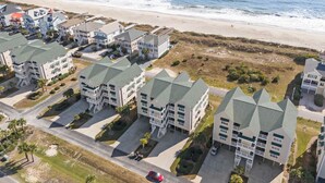 Un quartier résidentiel en bord de mer, composé de plusieurs immeubles à deux étages dotés de toits végétalisés et de balcons, avec une plage de sable fin et une vue dégagée sur l’océan.