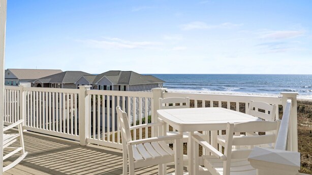 Une terrasse en bois avec des rampes blanches, une table et des chaises, donnant sur une plage avec des maisons en arrière-plan.