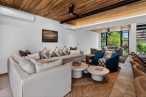 Una sala de estar moderna con un sofá, sillones y una mesa de centro. Hay unidades de aire acondicionado en la pared, un ventilador de techo y ventanas grandes con vistas a zonas verdes.