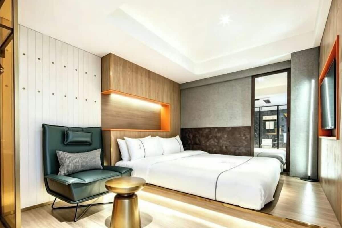 A modern hotel room with a large bed, a seating area with a chair and a small table, and a large window providing a view.