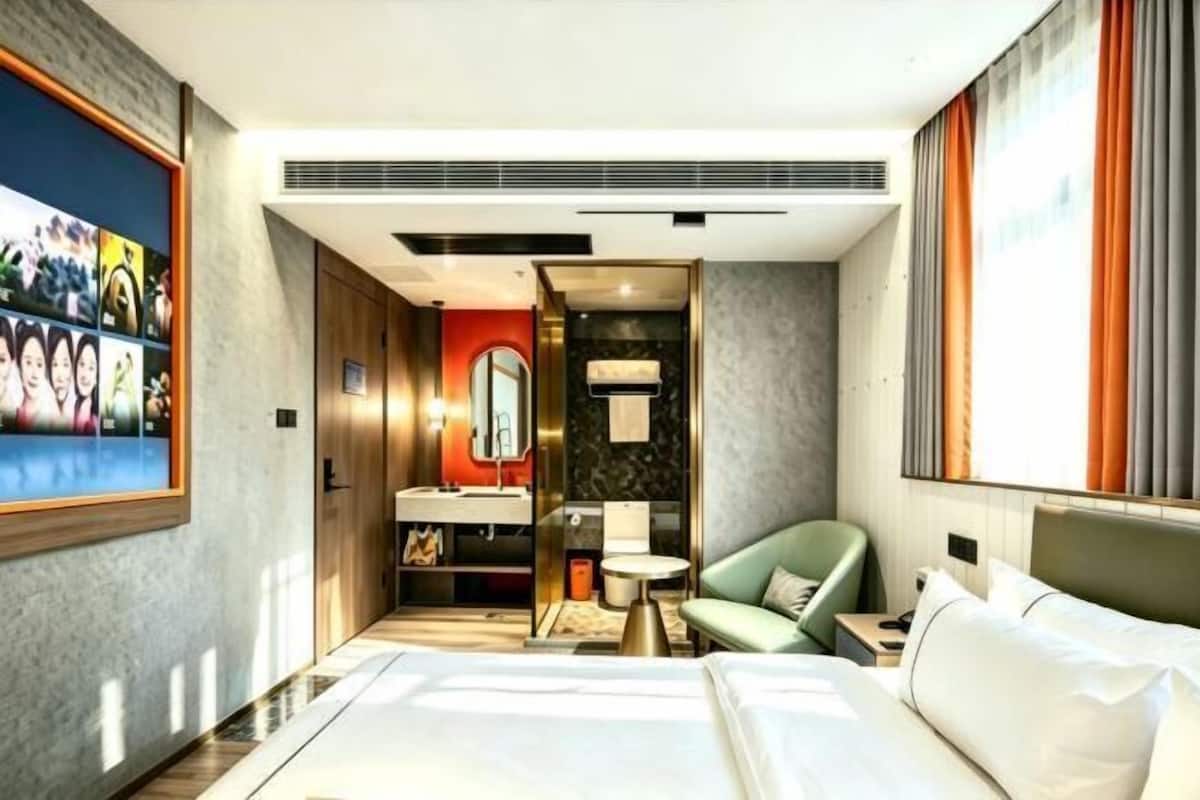 A hotel room with a bed, a green armchair, a small round table, a mirror, and a bathroom visible through an open door.