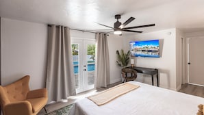 A bedroom with a bed, a ceiling fan, a TV on the wall, a desk, and a view of a pool through the door.