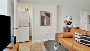A modern living room with a sofa, a coffee table, a TV, and a washing machine in the background. - Pet-Friendly 1-Bed, Walk to Fluor Field & Downtown (Greenville)