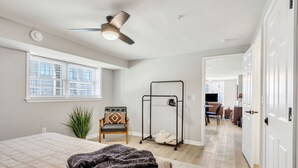 A bedroom with a bed, a ceiling fan, a window, a chair, a coat rack, and a hallway leading to a living area. - Pet-Friendly 1-Bed, Walk to Fluor Field & Downtown (Greenville)
