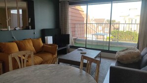 A living room with a sofa, a coffee table, and a TV. There is a window with a view of buildings outside.