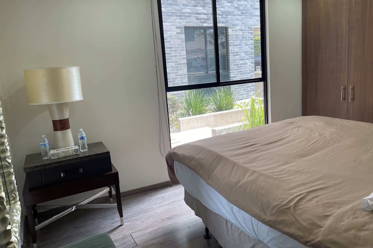 A bedroom with a bed, a nightstand with a lamp, two water bottles, and a window with a view of a brick building and greenery.