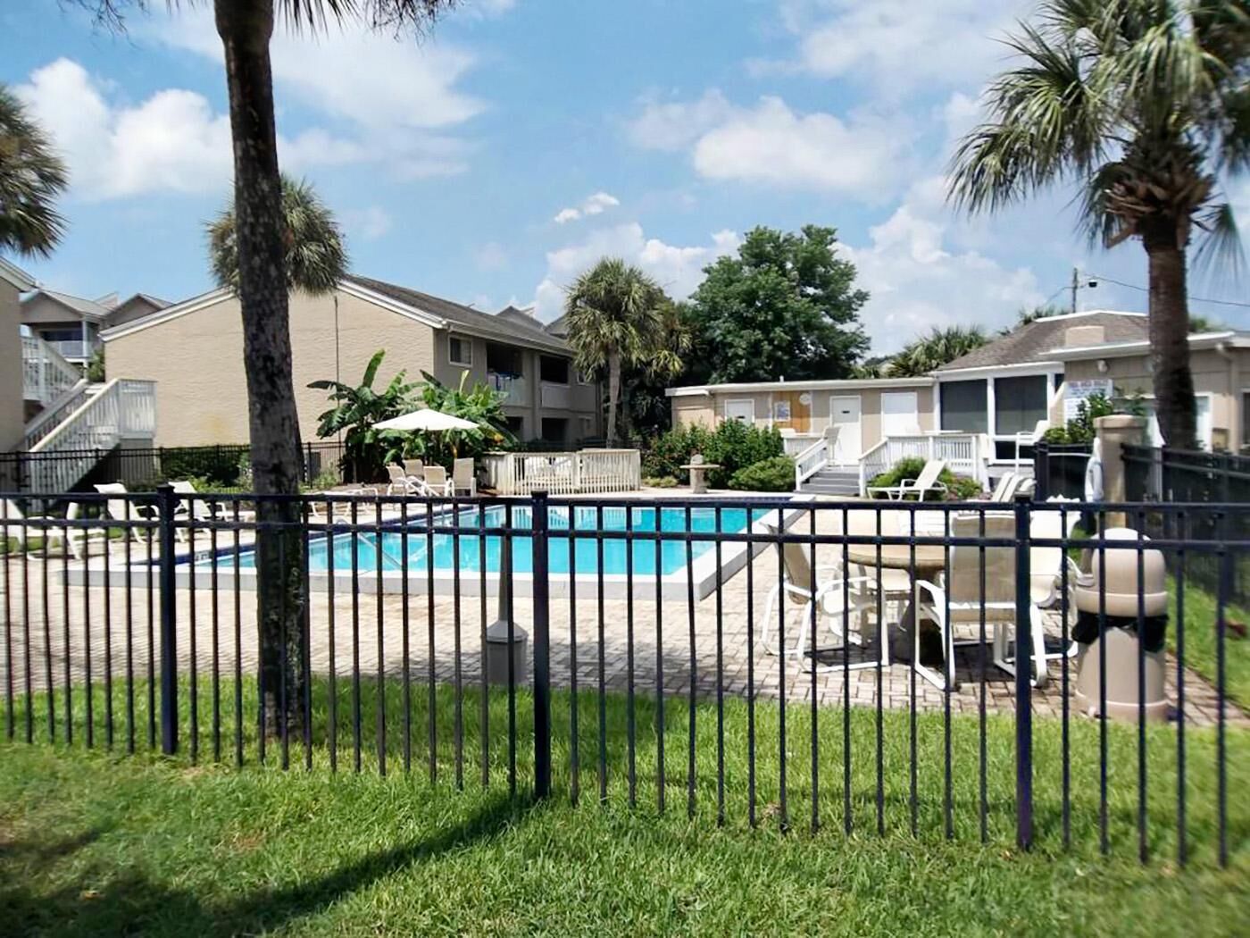 A swimming pool with lounge chairs and a fence, surrounded by palm trees and a building with balconies.