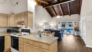 A spacious kitchen with wooden cabinets, a microwave, and a stove. There is a dining area with a table and chairs, and a living room with a sofa. - Chateau La Mer #7D (Destin)