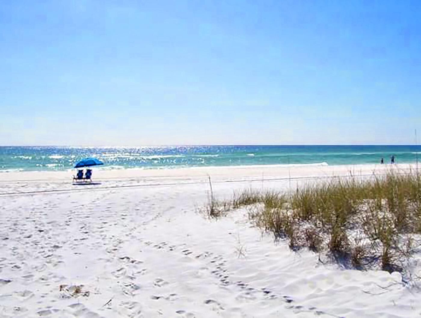 A sandy beach with a clear sky and ocean. There is a blue umbrella and a beach chair.