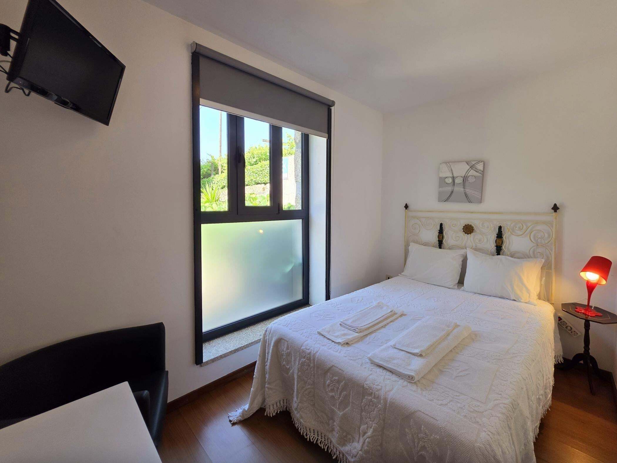 Un dormitorio con una cama, televisor montado en la pared, ventana con persianas y una lámpara de noche.
