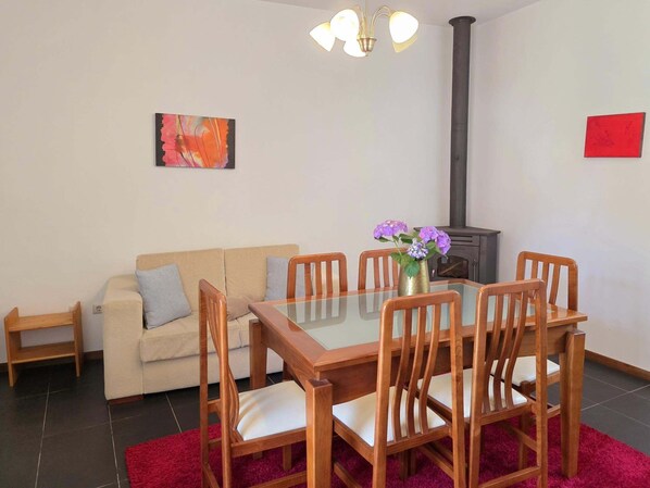 Una sala de estar con un sofá, una mesa de comedor con cubierta de vidrio, sillas de madera, un tapete rojo, un florero con flores, un cuadro y un estufa de leña.