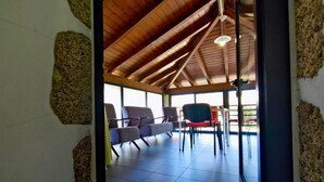 A covered outdoor seating area with chairs and tables, wooden ceiling beams, and large windows.