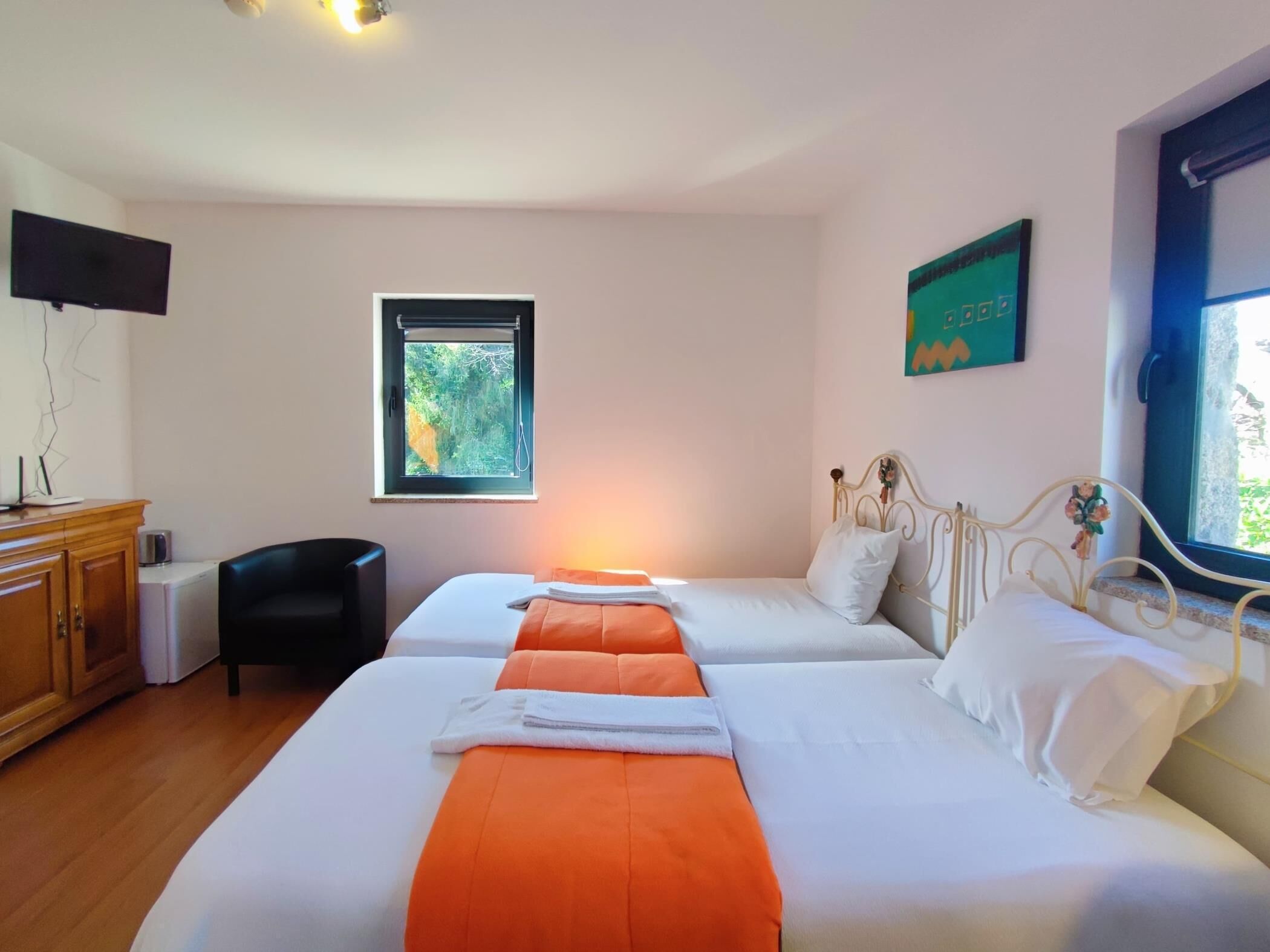 Habitación de hotel con dos camas, un armario de madera, televisor, ventana con vista a follaje y un cuadro en la pared.