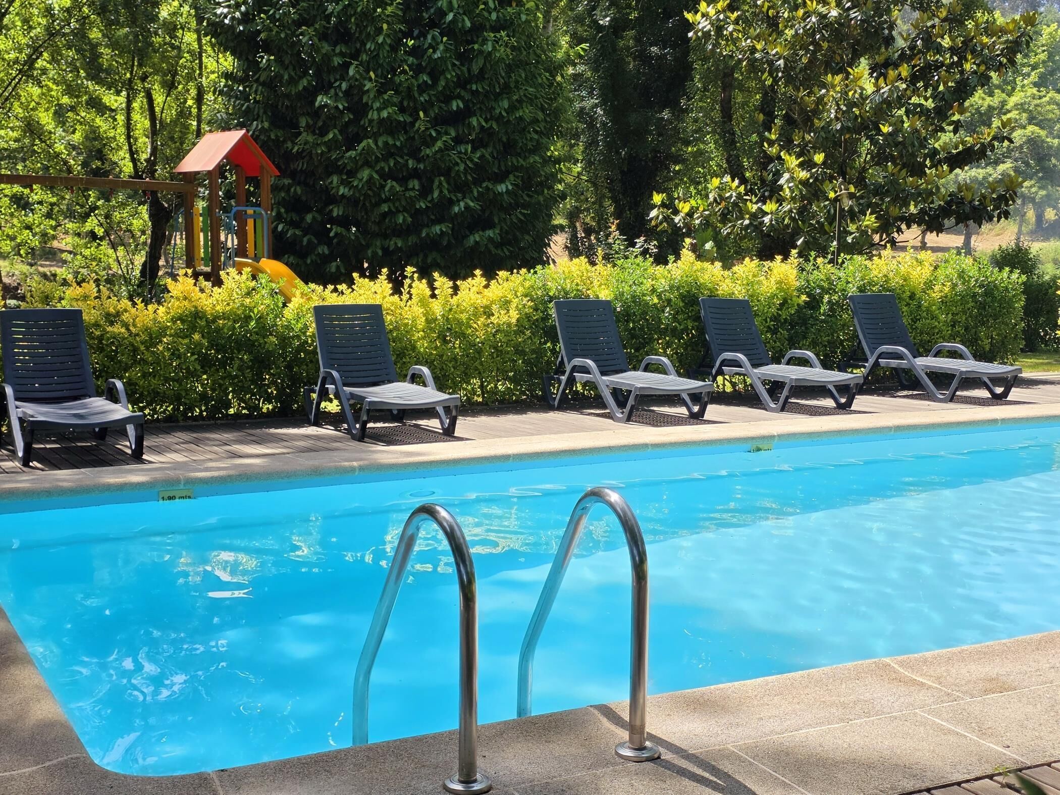 A swimming pool with a metal ladder, surrounded by lounge chairs and a small play structure.