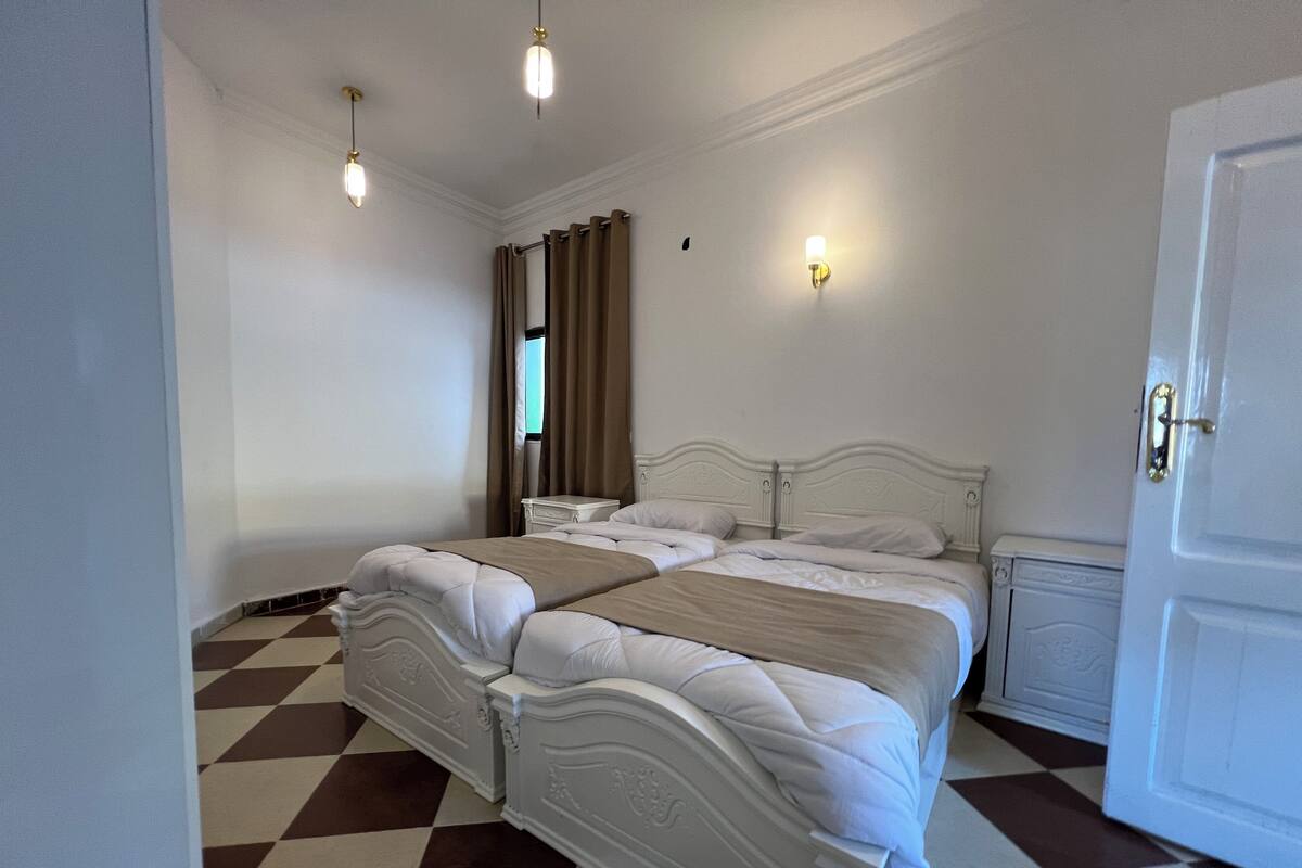 A hotel room with a checkered floor, a white bed with a headboard, two bedside tables, and a door.