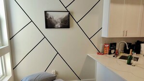 Une chambre compacte comprenant un lit, une petite table de chevet, une commode, une fenêtre et un coin cuisine avec des placards et un évier.