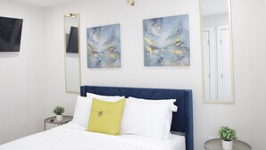 Une chambre à coucher comprenant un lit, deux tables de chevet, un grand miroir et trois tableaux abstraits accrochés au mur.