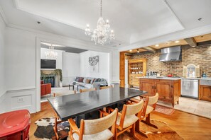 A spacious dining area with a long table and chairs, a chandelier, and a kitchen with brick walls and modern appliances.