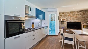 A modern kitchen with white cabinets, a built-in oven, and a dining area with a stone wall and a flat-screen TV. - Chic 3-bedroom apartment with AC, WiFi in charming Žrnovnica (Žrnovnica)