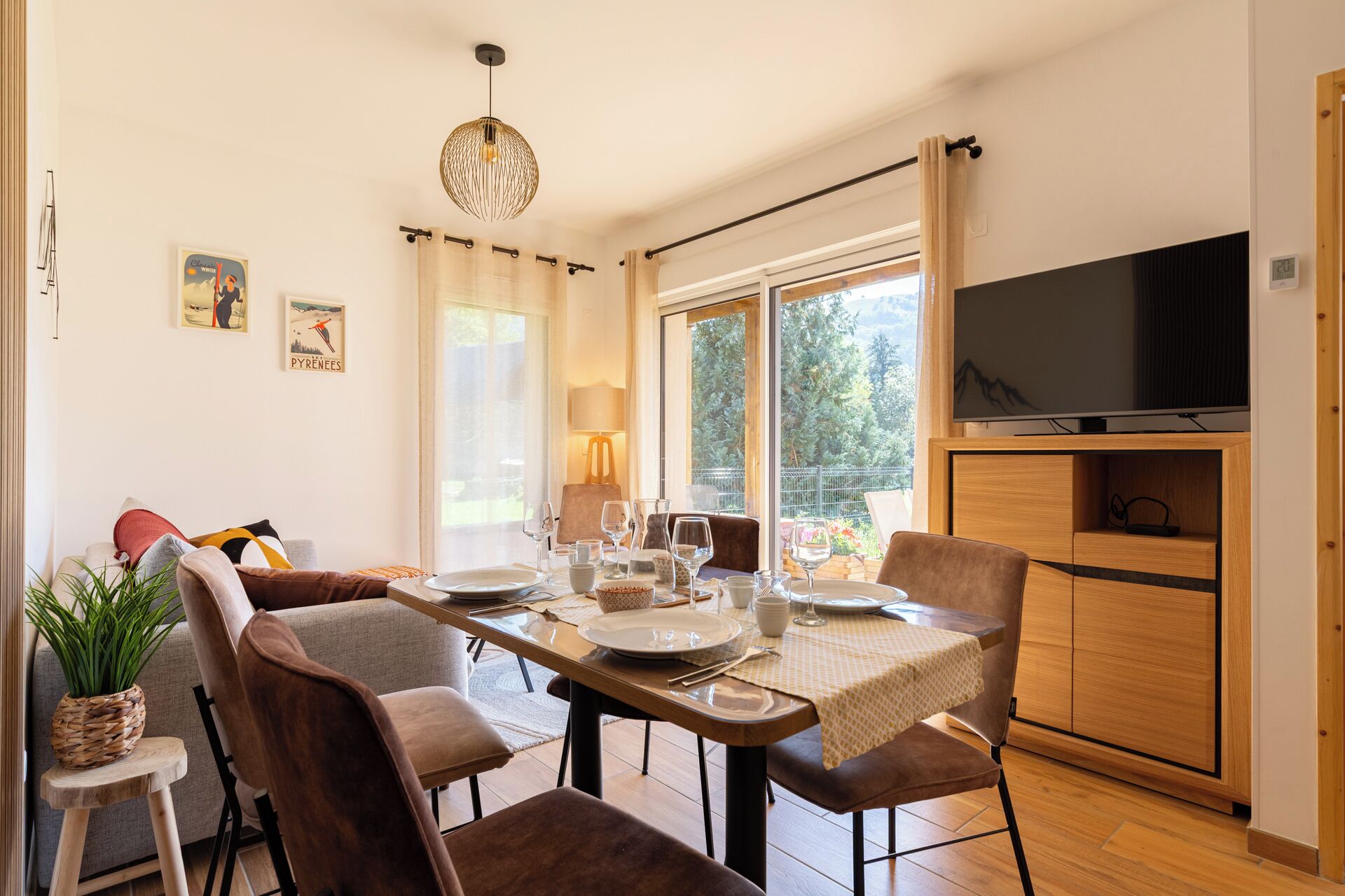 A dining area with a wooden table set for four, surrounded by chairs. A television is mounted on the wall to the right.