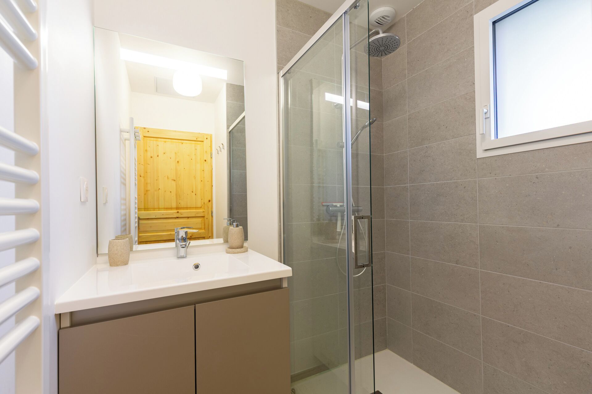 Un bany modern amb una cabina de dutxa de vidre, una porta de fusta i un moble de bany blanc amb mirall.