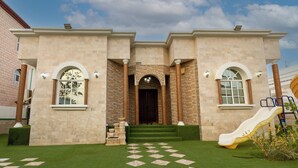 A two-story building with a central entrance, a brick facade, and a green lawn with a play area. - Bait Allubana Villa (Seeb)