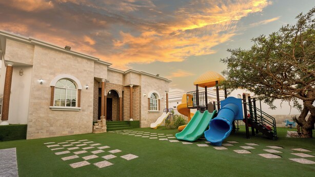 A playground with slides and a climbing structure, set against a building with a stone facade and arched windows. - Bait Allubana Villa (Seeb)
