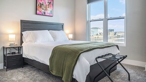 Una cama bien hecha con sábanas blancas, un plaid verde y una silla de metal negro al pie. Un mesita de noche con una lámpara y una pequeña mesa están junto a la cama. Un ventanal grande ofrece vistas a edificios y un cielo despejado.