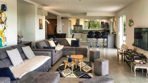 Ein modernes Wohnzimmer mit einer grauen Sofagarnitur, einem Couchtisch und Hocker. Im Hintergrund ist eine Küche mit Barhocker zu sehen.