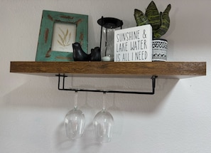 A shelf with a framed botanical print, a black decorative object, a sign with text, a patterned pot, and two wine glasses hanging below.
