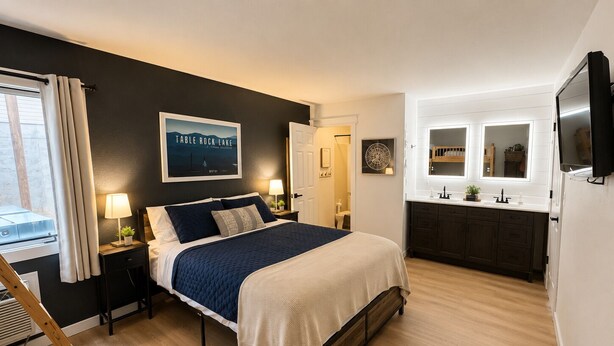 A bedroom with a bed, bedside tables, a nightstand, a window with curtains, a wall-mounted TV, a kitchen area with cabinets, a countertop, and a wall-mounted mirror.
